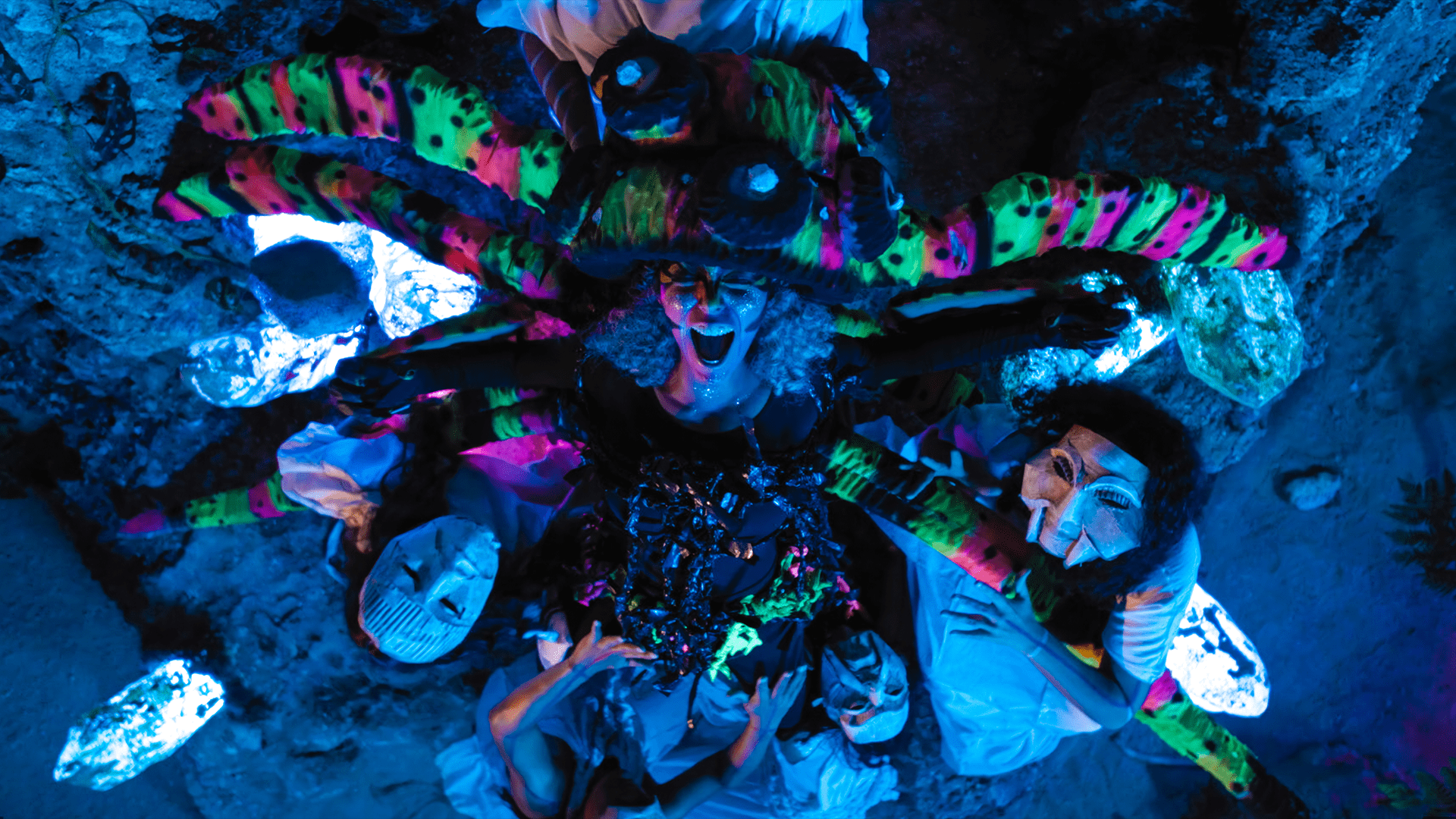 Clean, high-resolution overhead cinematic still from the movie Matininó. In a dimly lit cave illuminated by glowing blue rocks, a central female performer in neon green, pink, and black spider-like costuming screams upward. Around her, performers in white dresses wear dramatic, stylized theatrical masks.