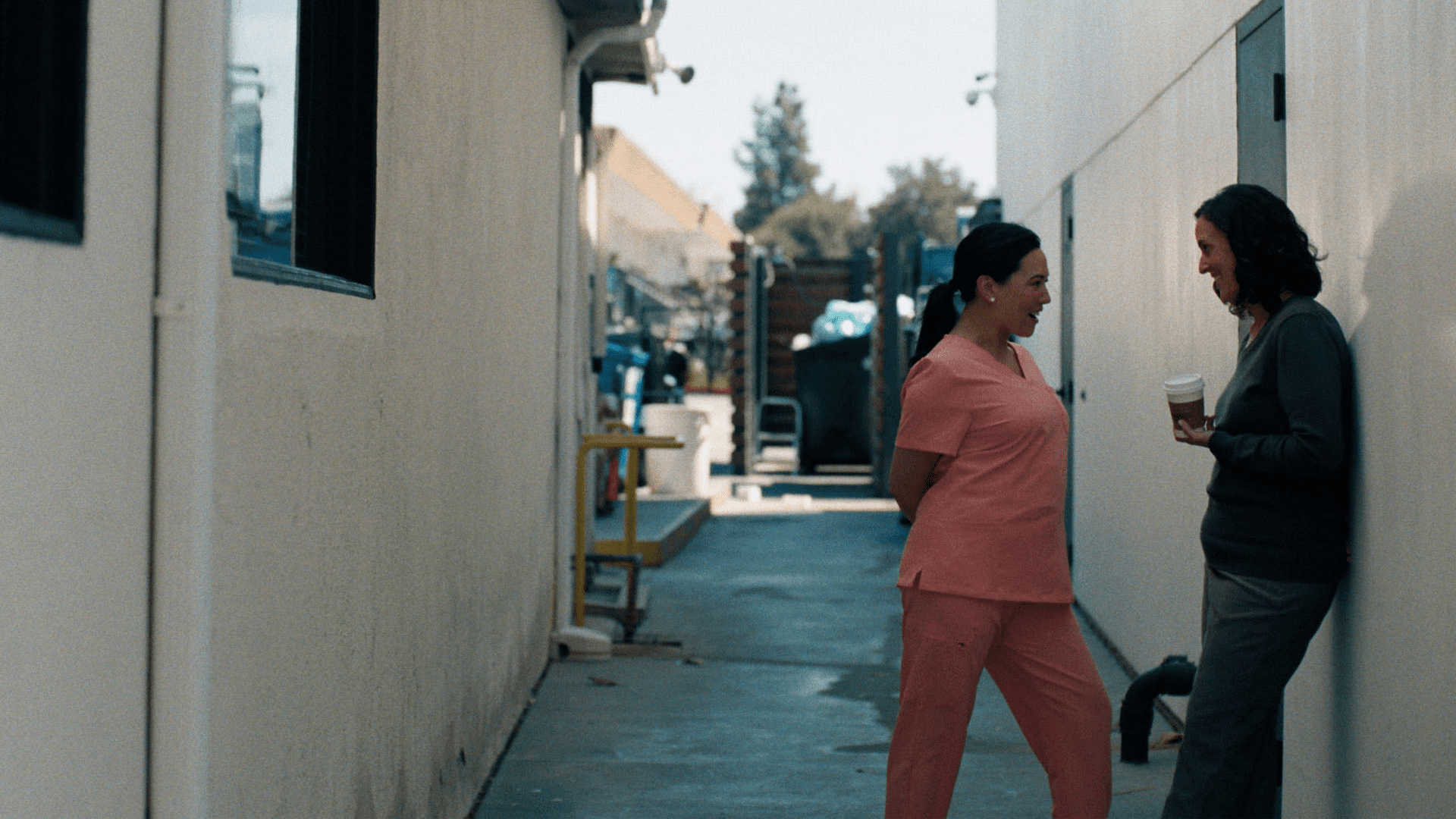 Clean, high-resolution 16x9 cinematic still from the dark comedy film She Keeps Me Young. Two women stand chatting in a narrow, outdoor alleyway between white commercial buildings. On the left, a woman with dark hair tied back in a ponytail stands in pink medical scrubs. On the right, a woman in a dark long-sleeve shirt and grey pants leans against the wall while holding a disposable coffee cup.