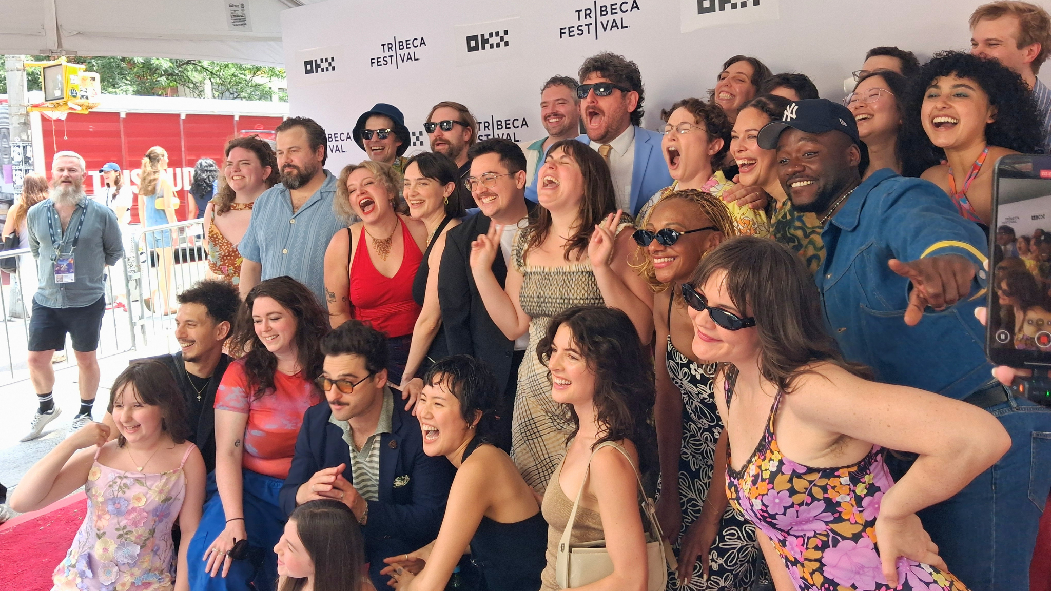 A large ensemble cast and crew pose together playfully, laughing and shouting on the outdoor red carpet under a white tent for the Tribeca Festival world premiere of Human Theories. The background features a step-and-repeat wall displaying the Tribeca Festival and OKX logos.