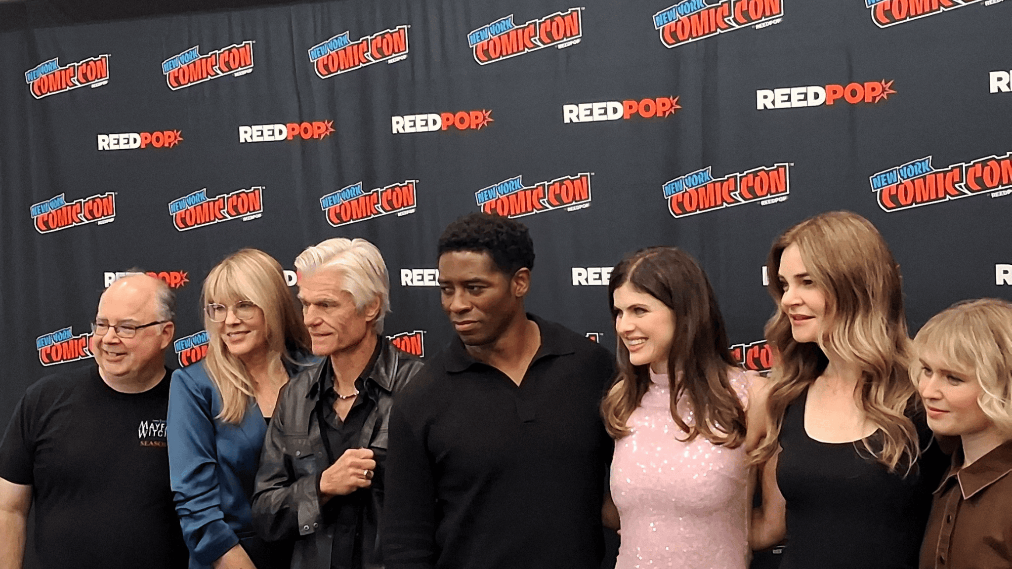 Medium shot of the Mayfair Witches: Salem Coven cast and executive producers posing closely together on the New York Comic Con 2026 press line. Standing left to right in front of a black backdrop with repeating New York Comic Con and ReedPop logos are co-showrunner Thomas Schnauz, co-showrunner Esta Spalding, Harry Hamlin, Tongayi Chirisa, lead actor Alexandra Daddario smiling in a pink sequined dress, Betsy Brandt, and Eliza Scanlen.